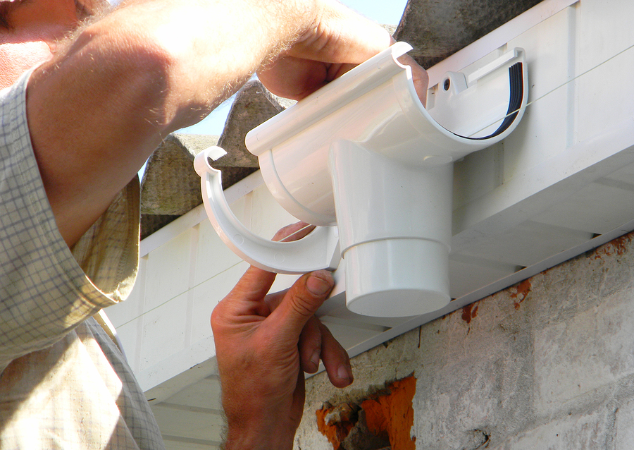 contractor installing plastic roof gutter holder for downspout drain pipe. plastic roof guttering, rain guttering & drainage by handyman hands.